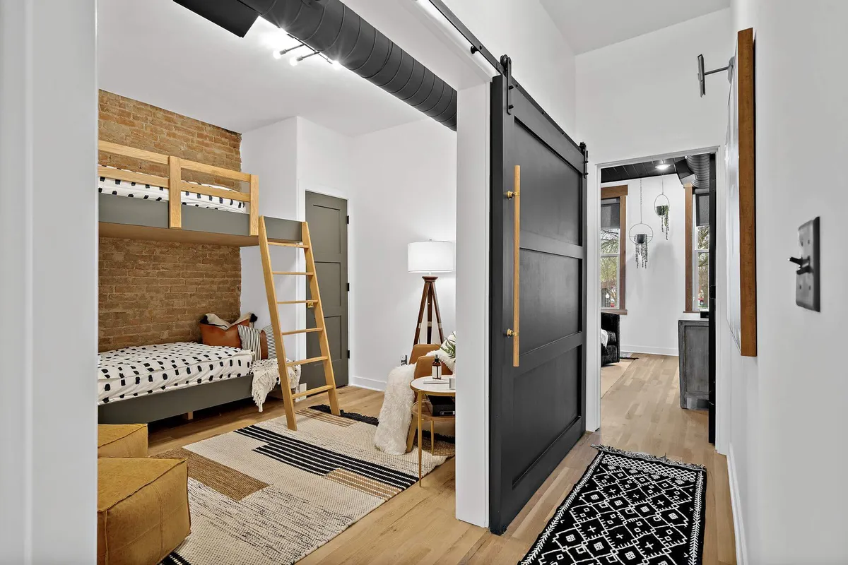A cozy room featuring built-in bunk beds with a wooden ladder, a sliding barn door, a textured area rug, and a warm lighting fixture. The space is inviting and ideal for groups to gather for games, reading, or winding down together, with playful touches that suggest shared experiences.