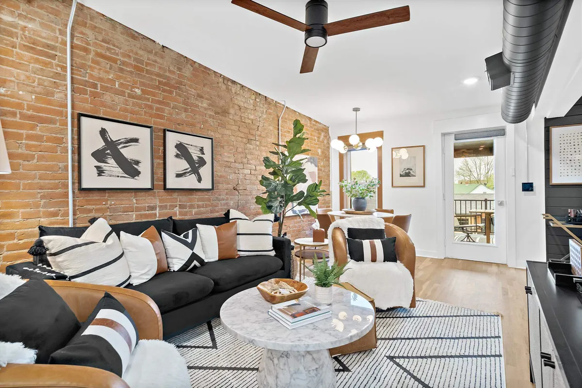 A cozy living area featuring an exposed brick wall, comfortable black sofa with decorative pillows, a round marble coffee table, and lush indoor plants. The setting suggests a space perfect for relaxing, socializing, and enjoying indoor comfort.