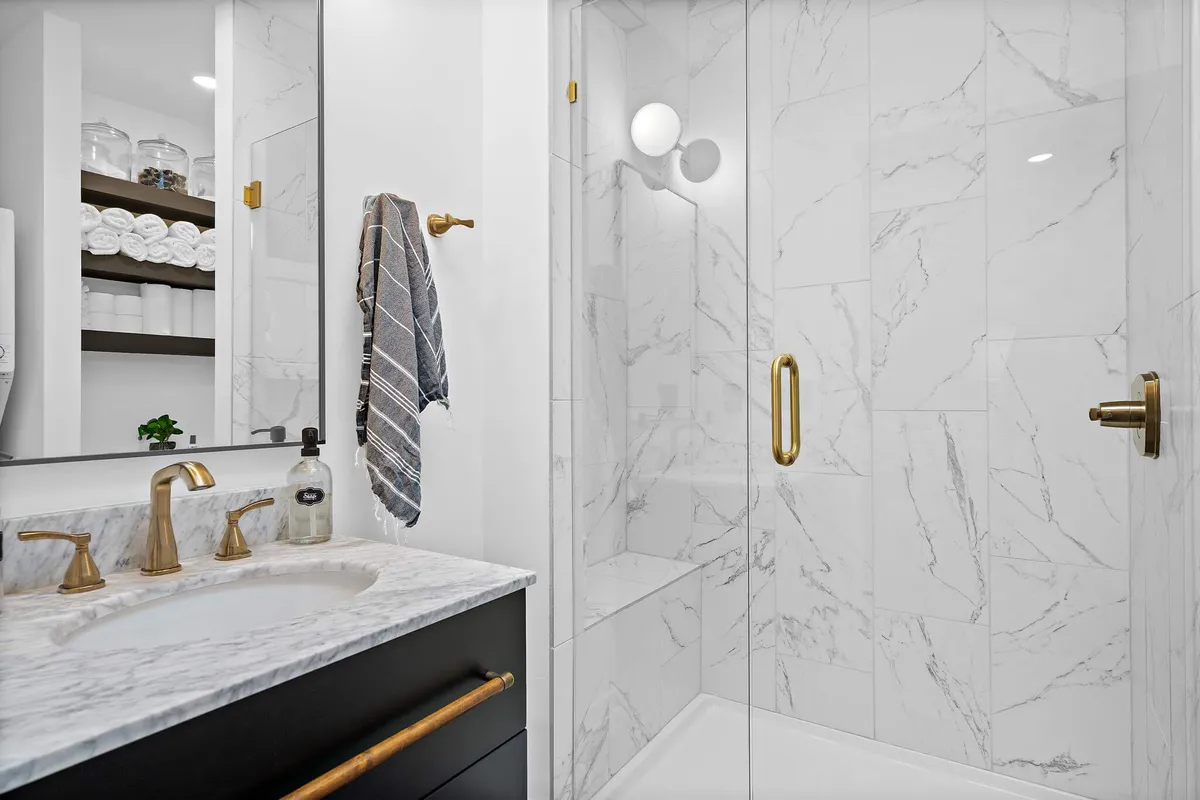A modern bathroom featuring a marble countertop, gold fixtures, a glass shower enclosure, and neatly arranged towels. The design is clean and serene, offering a spa-like atmosphere for self-care and relaxation.