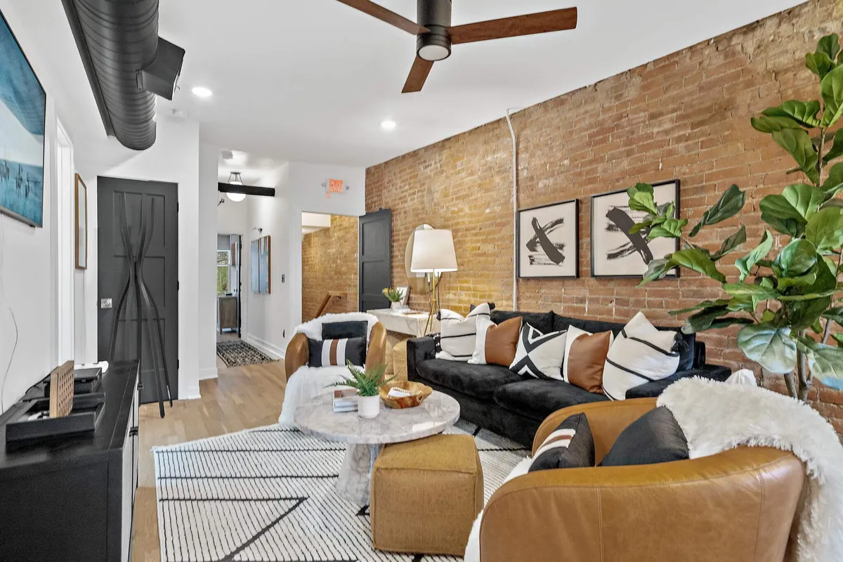 An inviting living room with a black couch adorned with colorful pillows, a round marble coffee table, a large indoor plant, and framed artwork. Wooden furniture accents add warmth and personality to the space.