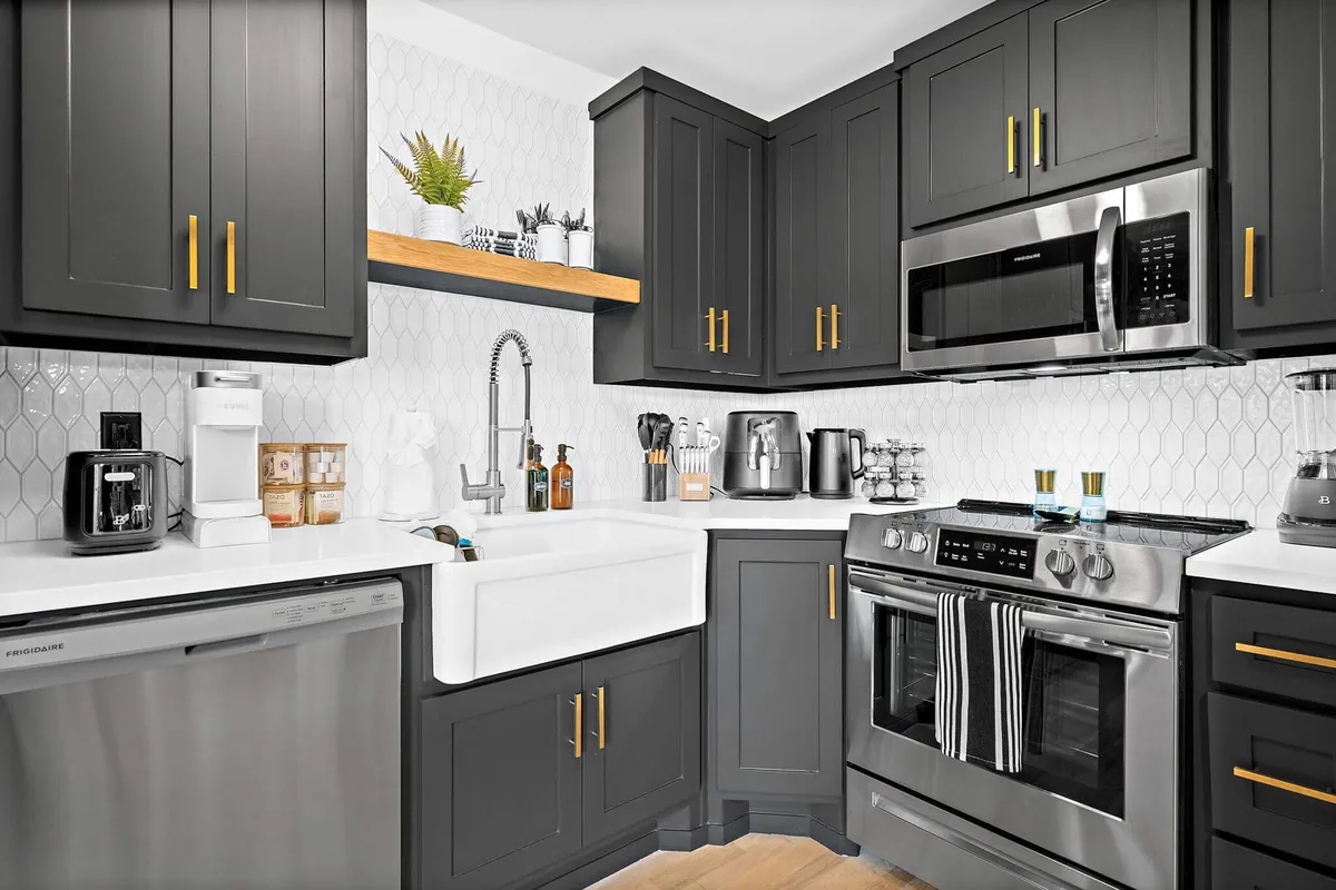A stylish, open kitchen with black cabinets, a white farmhouse sink, stainless steel appliances, and a geometric backsplash. The area is arranged for social cooking, entertaining, and gathering, offering a functional and social focal point for the home.