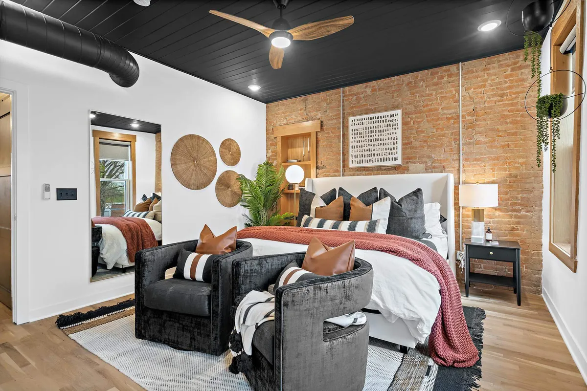 An inviting bedroom with a bed covered in decorative pillows, a mirrored wardrobe for practical storage, wooden wall art, a potted plant, and a black ceiling fan. The space is bathed in soft morning light, creating a cozy yet stylish environment.