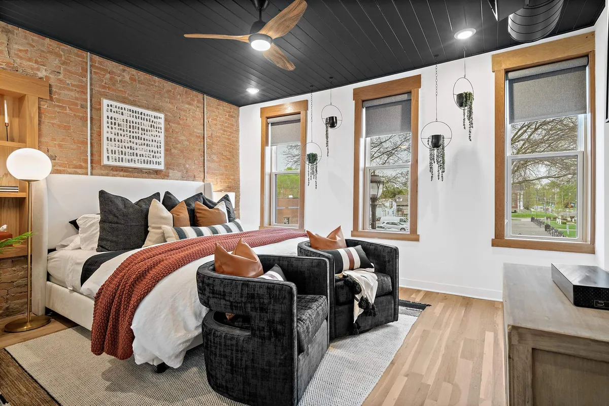 A welcoming bedroom featuring a bed adorned with decorative pillows, flanked by black armchairs for reading or relaxing. Hanging plants and large windows allow plenty of natural light, enhancing the inviting and restful atmosphere.