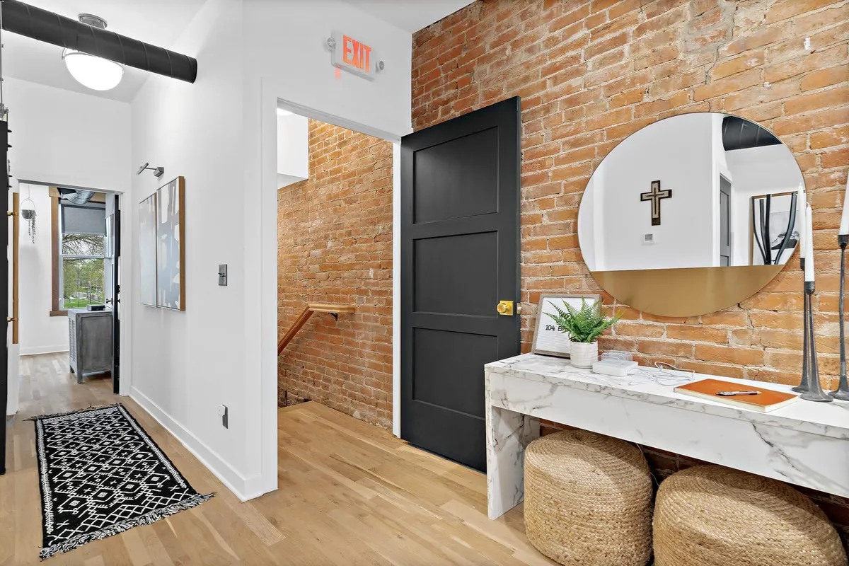 This image showcases the entryway with its exposed brick wall, sleek black door, round mirror, marble console table, and woven stools. The ambiance is inviting and modern, setting the tone for the home's urban loft style.