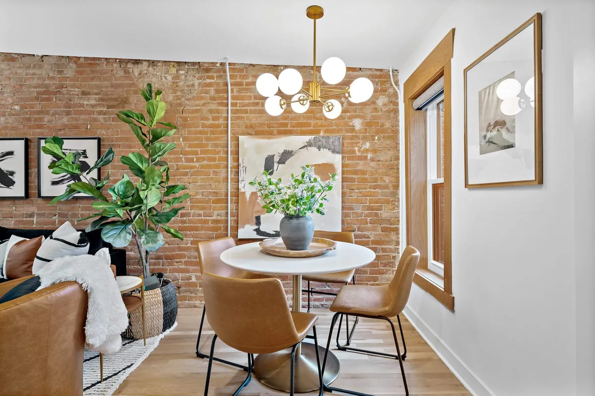 A modern dining area with a brick wall, elegant chandelier, dining table, potted plant, and artwork. The mood is cozy yet stylish, evoking intimate meals and social gatherings.