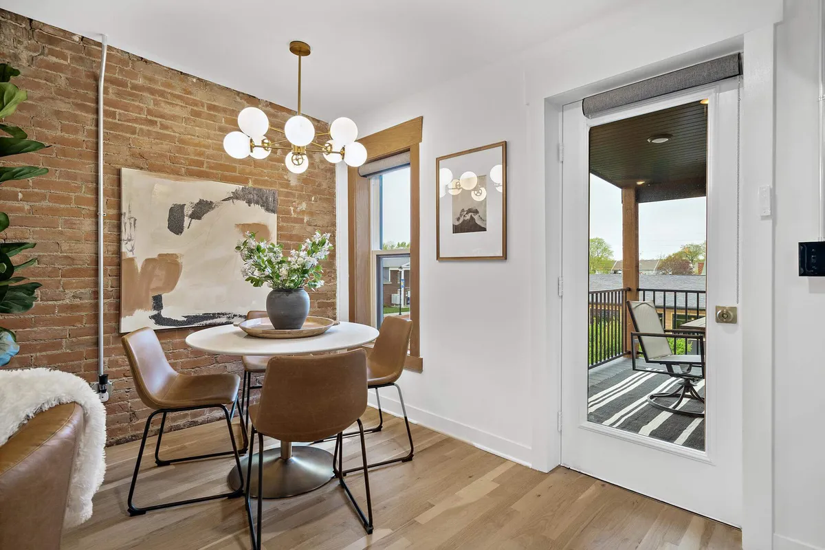 A warm dining area with a large wooden table, designer lighting, brick wall art, and a lush green plant. The space is adjacent to a door that leads to a balcony, inviting morning light and offering a cozy spot for meals and gatherings.