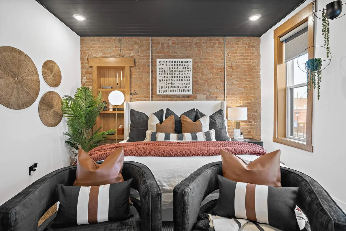 A welcoming bedroom featuring a bed with decorative pillows, an exposed brick wall, hanging plants, and artwork. The space is cozy yet stylish, reflecting the comfort and charm of home after a day exploring Kansas City.